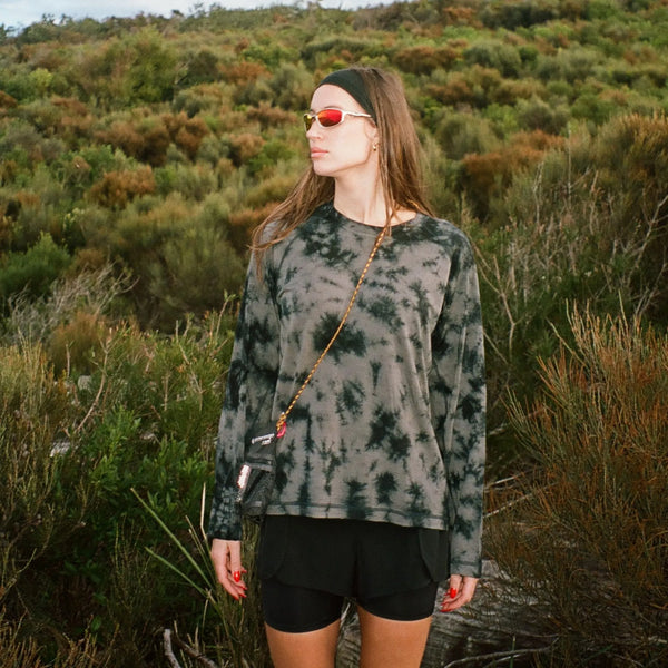 Woman wearing a tie-dye shirt and sunglasses standing in a natural landscape.