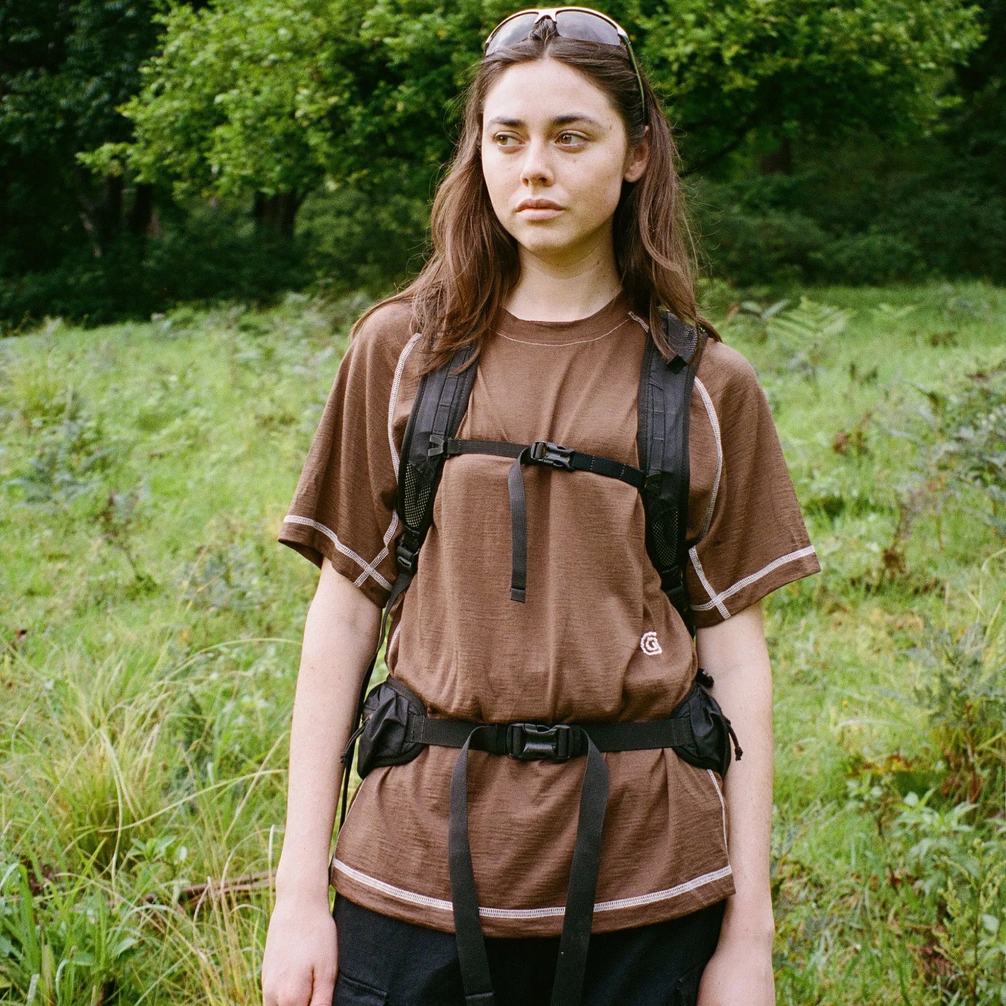 Person wearing a brown t-shirt and black backpack in a grassy outdoor setting