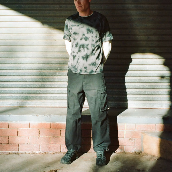 Person wearing a tie-dye shirt and cargo pants standing against a metal shutter.