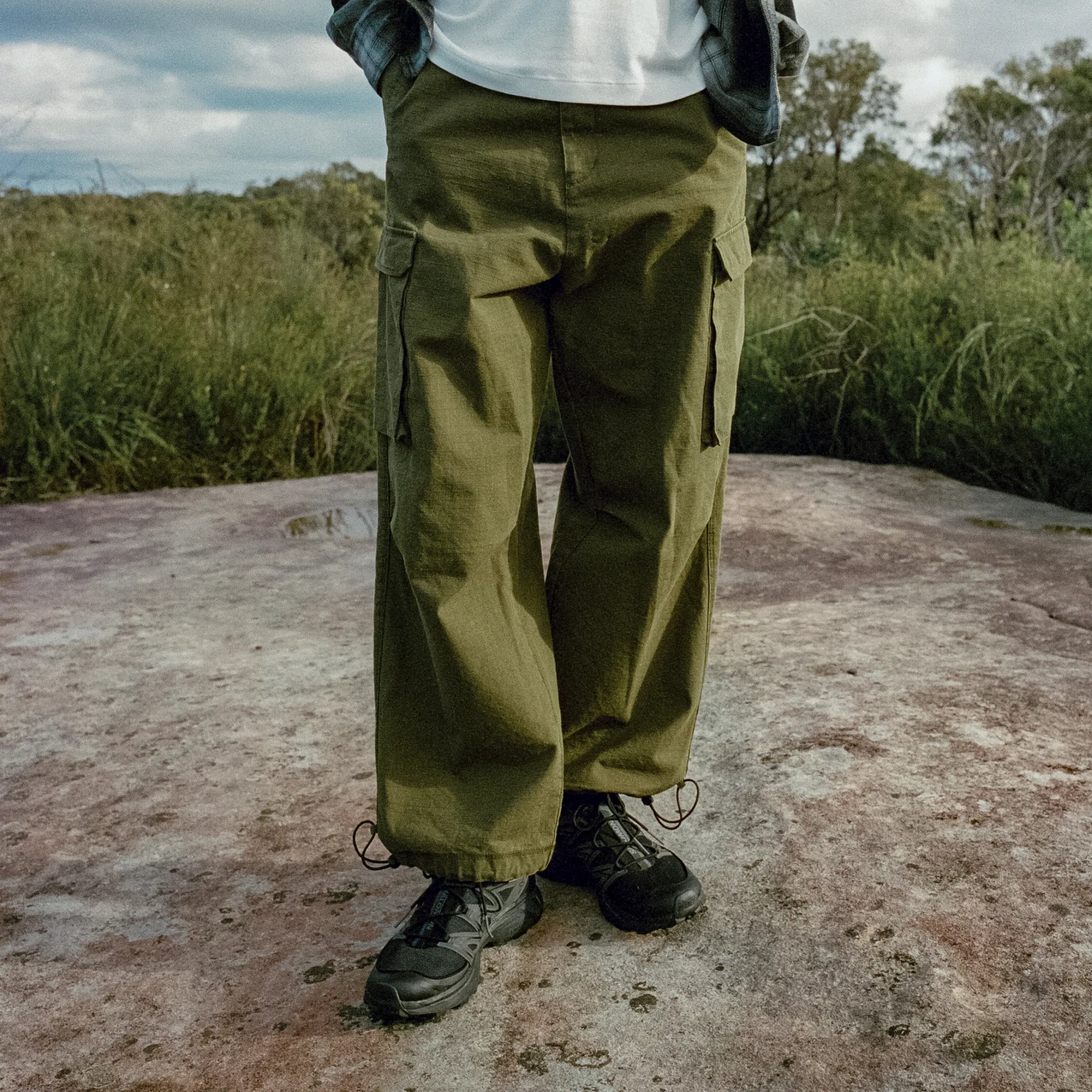 Person wearing green cargo pants and black shoes standing on a rocky surface with grass and trees in the background.