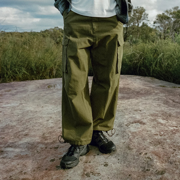 Person wearing green cargo pants and black shoes standing on a rocky surface with grass and trees in the background.