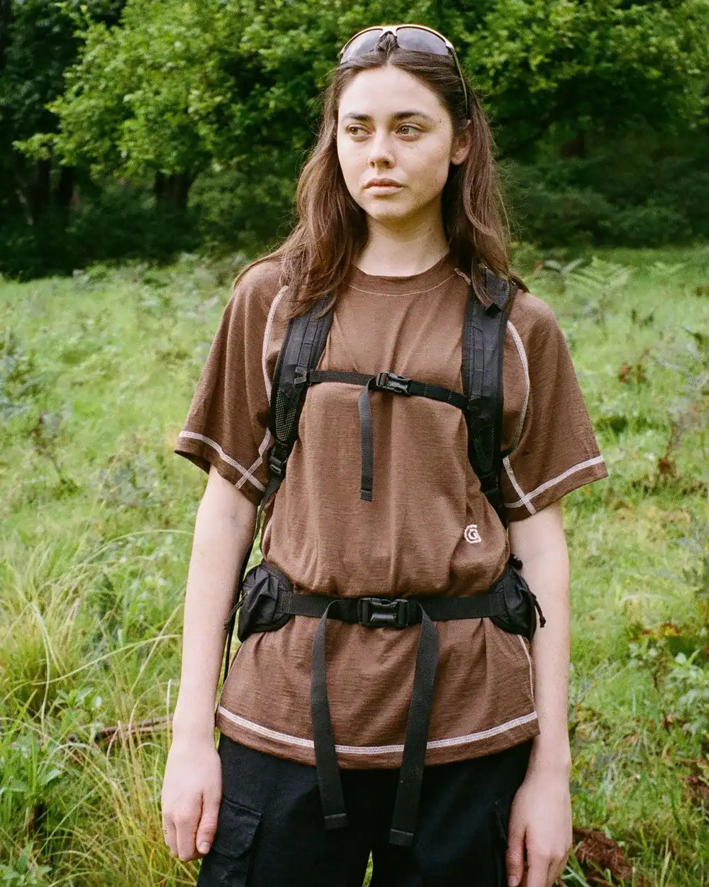 Person wearing a brown t-shirt and black backpack in a grassy outdoor setting